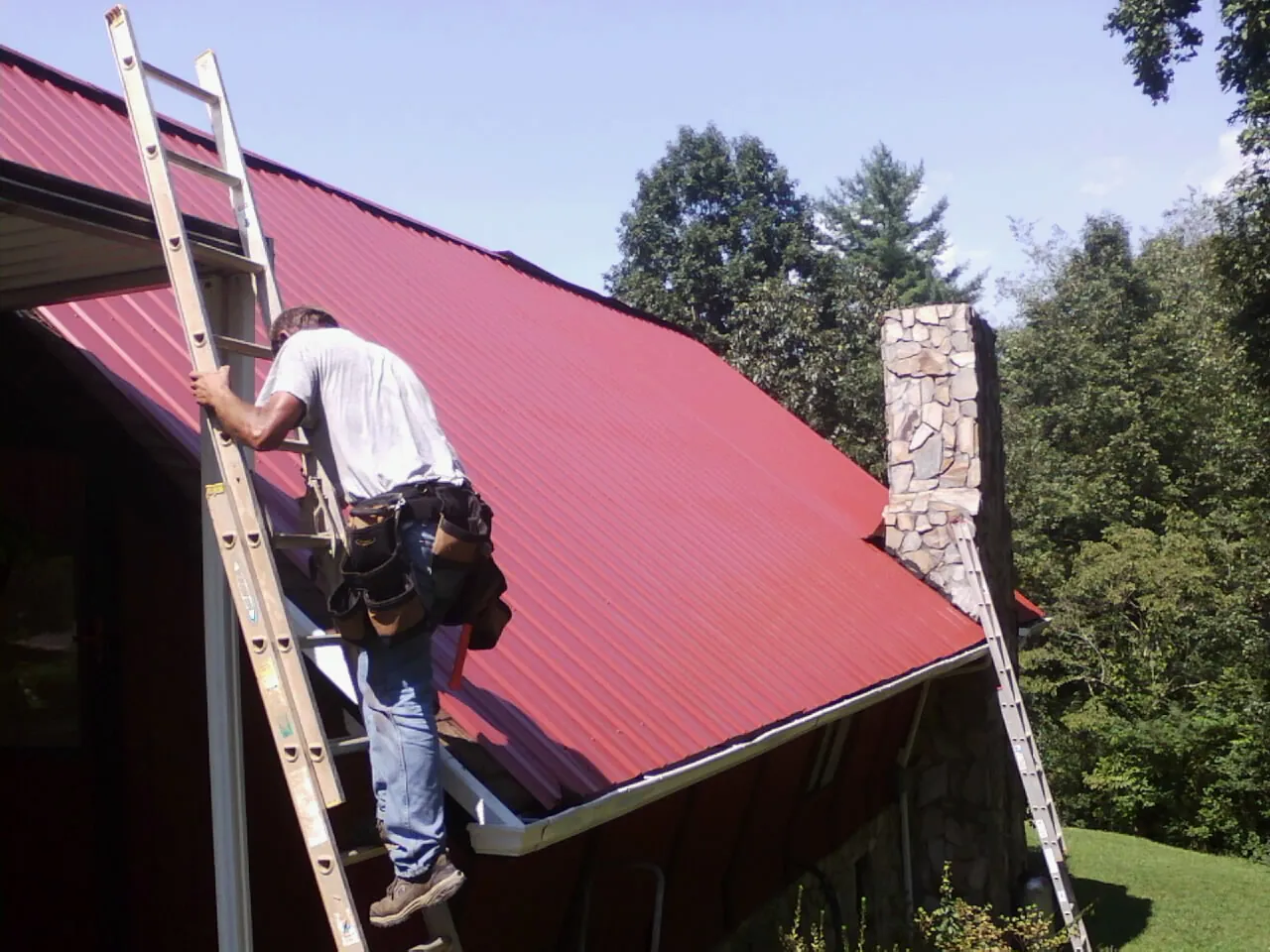 Roofer on a ladder beside a red metal roof and stone chimney — Soffit & Fascia Repair by Roofing Brothers in Como